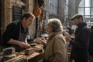 Inside a covered market in Quimper, a charcutier converses with an elderly woman at his stall while another customer selects artisan charcuterie, bathed in soft natural morning light.