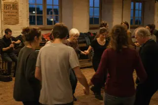 Inside a village hall in Finistère at dusk, people of various ages dance in a circle, motion slightly blurred, with coats and boots on benches, soft natural light and an authentic, understated atmosphere.