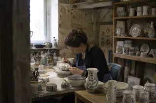 Artisan woman painting ceramic in a rustic faience workshop in Quimper, natural morning light casting soft shadows