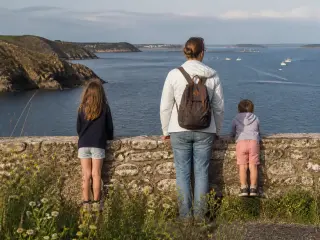 Two children and an adult pausing at a stone wall above Douarnenez bay, with soft late afternoon light and wildflowers in the foreground, looking out over the sea and distant boats.
